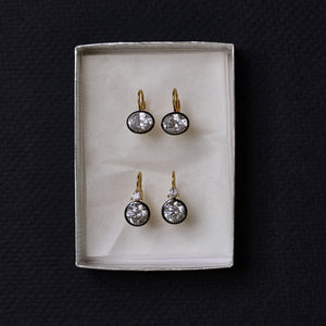 Top-down view of rhodium double bezel drop earrings showcasing round bezel settings in a white jewelry box on a dark background
