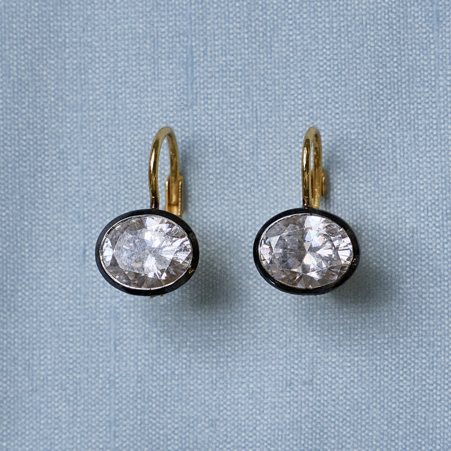Front view of Rhodium Oval Bezel Drop Earrings showcasing oval stones in dark rhodium bezels on light blue fabric