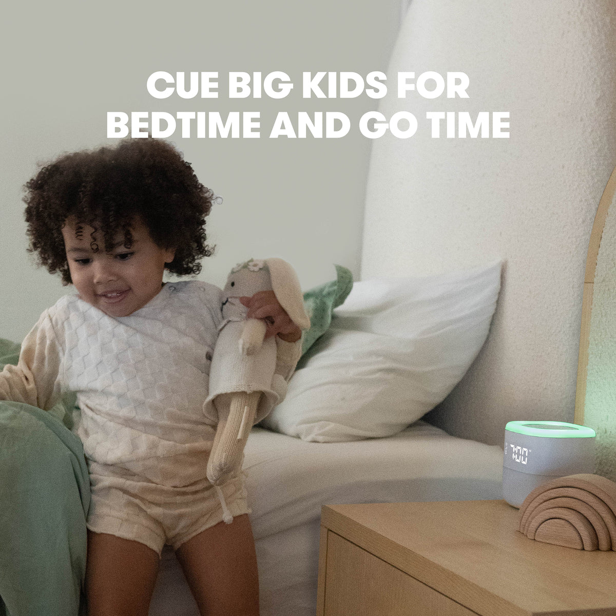 Toddler holding a stuffed rabbit on a bed next to a nightstand with a lit 3-in-1 sound machine showing a green wake light; overlay text CUE BIG KIDS FOR BEDTIME AND GO TIME