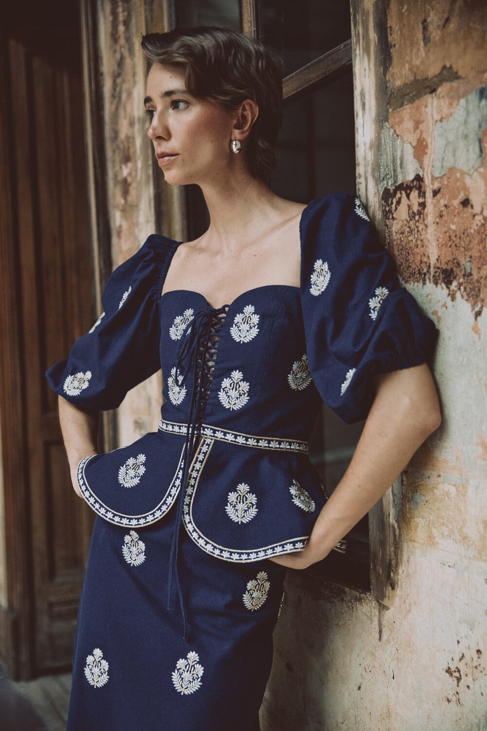 Three quarter view of Mitra navy lace front bustier and skirt with white embroidery, lace up front, puff sleeves, peplum waist