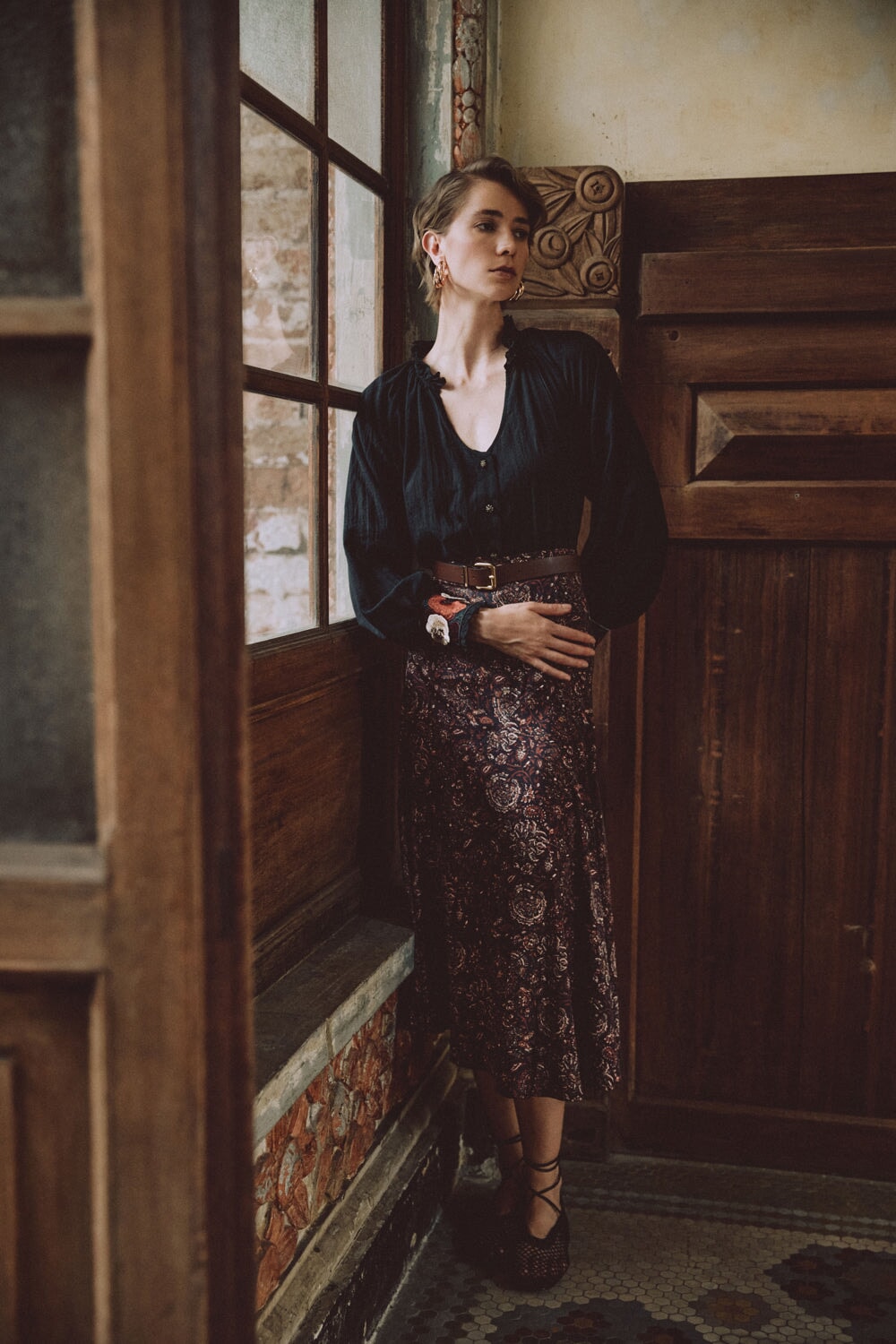 Model leaning against a window wearing Zoya printed silk brown skirt with belted waist in a vintage wood-paneled room