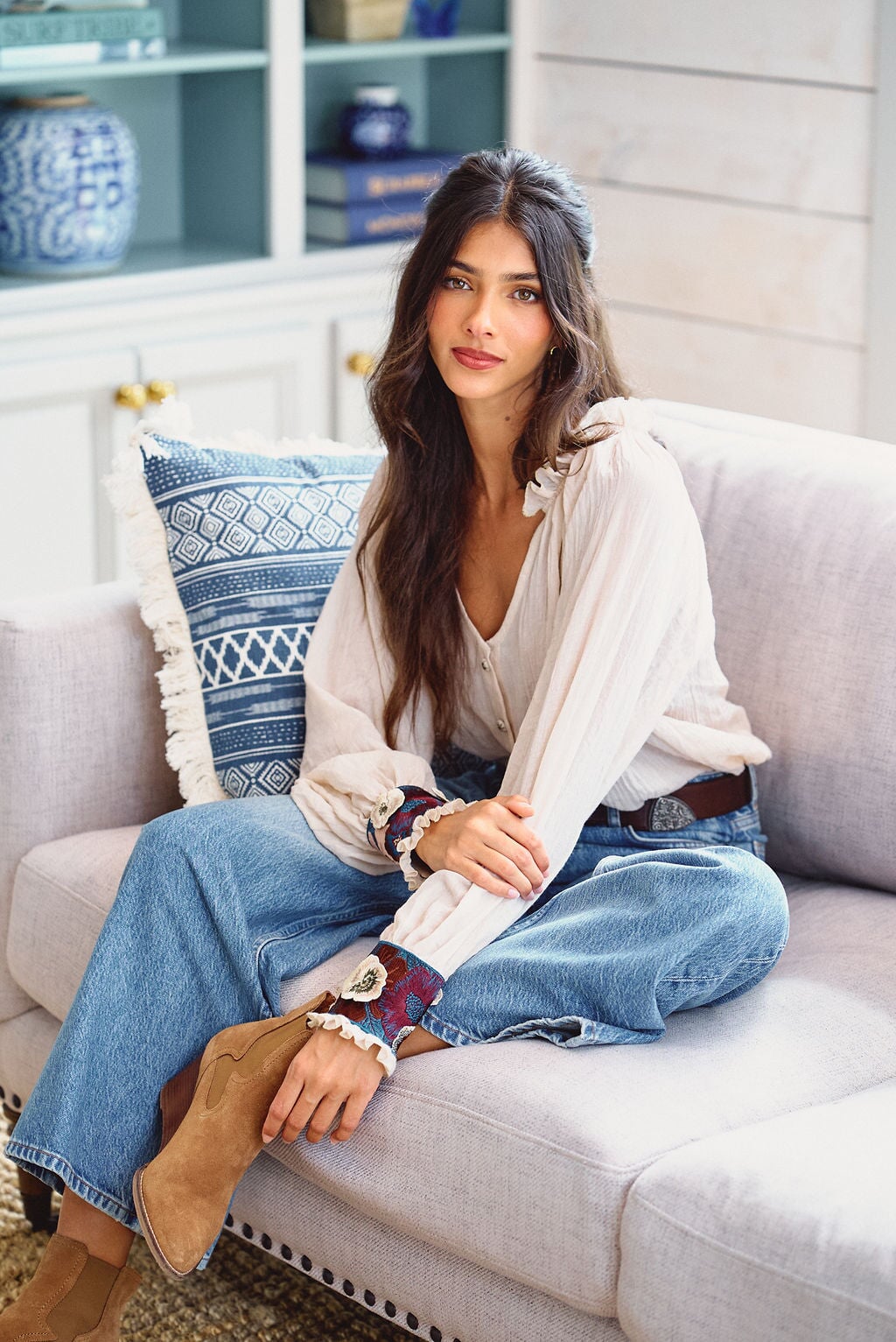 Front view of Pansy Blouse in Creme showcasing embroidered floral cuffs and billowy sleeves with a seated model on a sofa