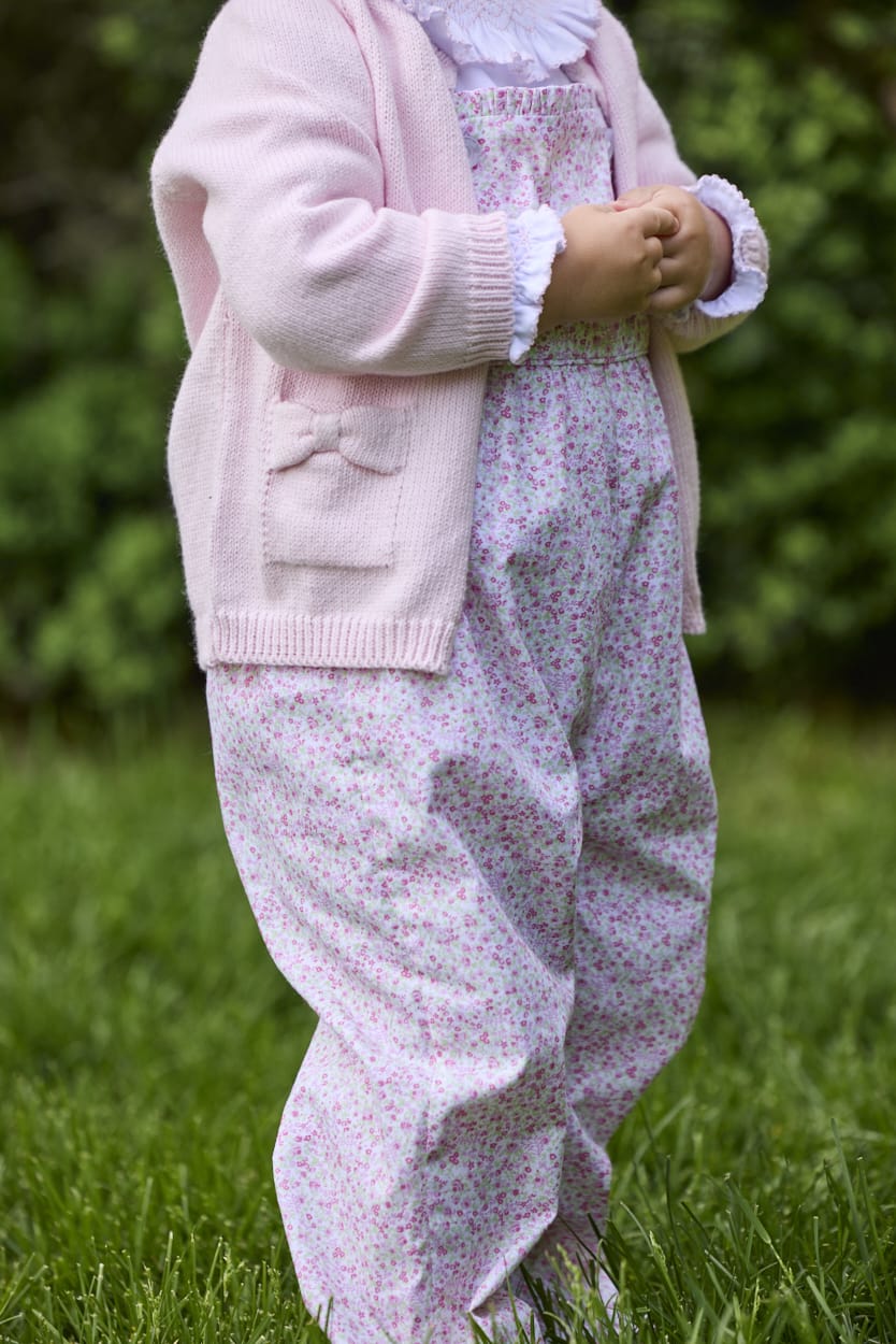 Little English traditional children's clothing.  Light pink and green floral overall with ruffle detailing layered over white knit smocked Madeline Blouse in light pink. Light pink cotton bow pocket cardigan added on top.