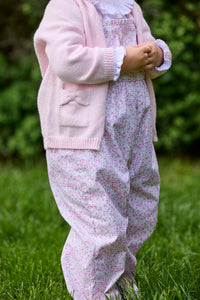 Little English traditional children's clothing.  Light pink and green floral overall with ruffle detailing layered over white knit smocked Madeline Blouse in light pink. Light pink cotton bow pocket cardigan added on top.