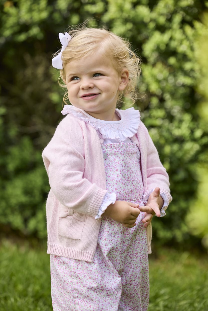 Little English traditional children's clothing.  Light pink and green floral overall with ruffle detailing layered over white knit smocked Madeline Blouse in light pink. Light pink cotton bow pocket cardigan added on top.