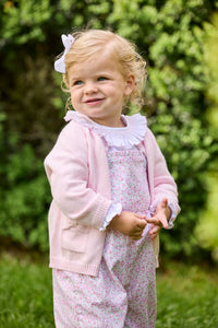 Little English traditional children's clothing.  Light pink and green floral overall with ruffle detailing layered over white knit smocked Madeline Blouse in light pink. Light pink cotton bow pocket cardigan added on top.