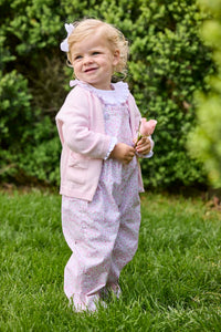 Little English traditional children's clothing.  Light pink and green floral overall with ruffle detailing layered over white knit smocked Madeline Blouse in light pink. Light pink cotton bow pocket cardigan added on top.