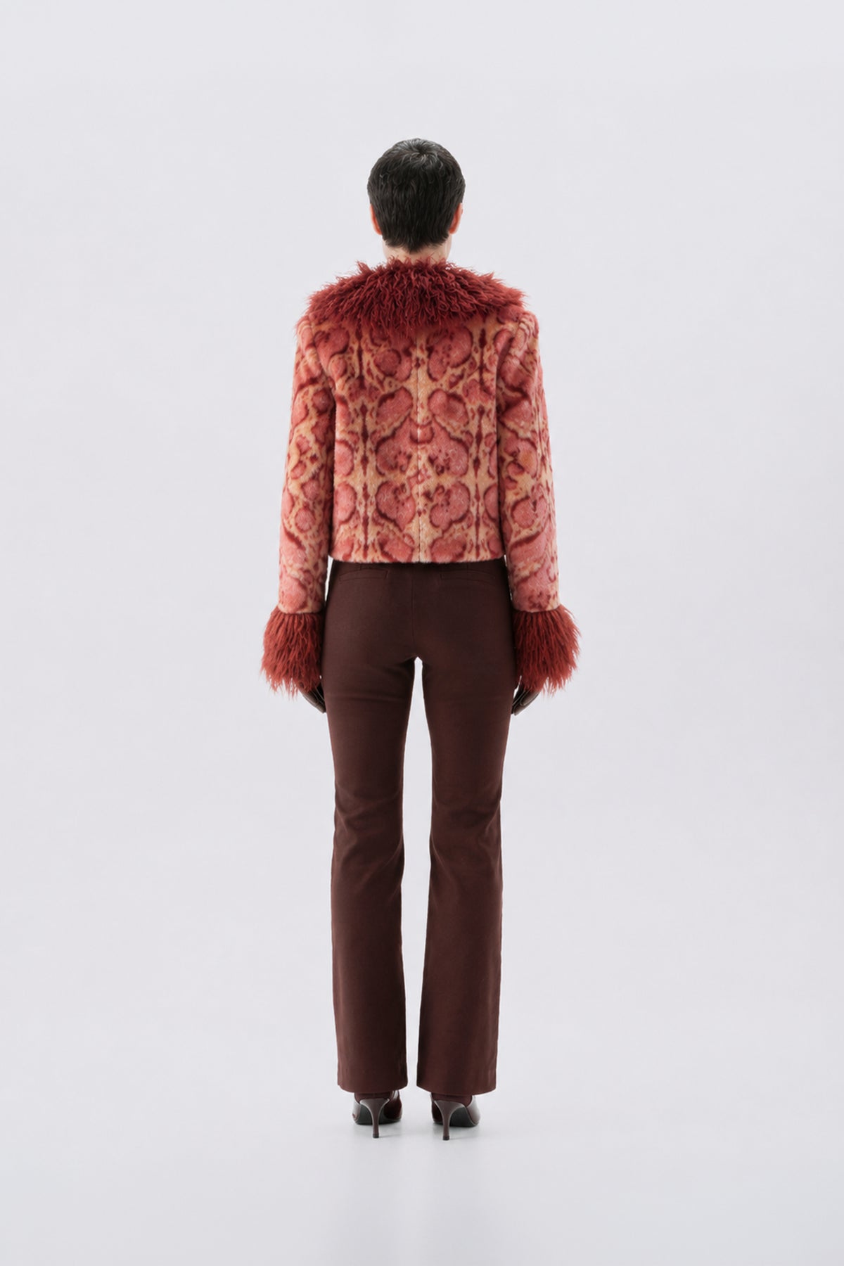 A model shown from the back wearing a cropped, patterned pink jacket with reddish-brown faux fur trim at the collar and cuffs, paired with dark brown trousers and black heels, against a light gray studio background.