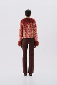 A model shown from the back wearing a cropped, patterned pink jacket with reddish-brown faux fur trim at the collar and cuffs, paired with dark brown trousers and black heels, against a light gray studio background.