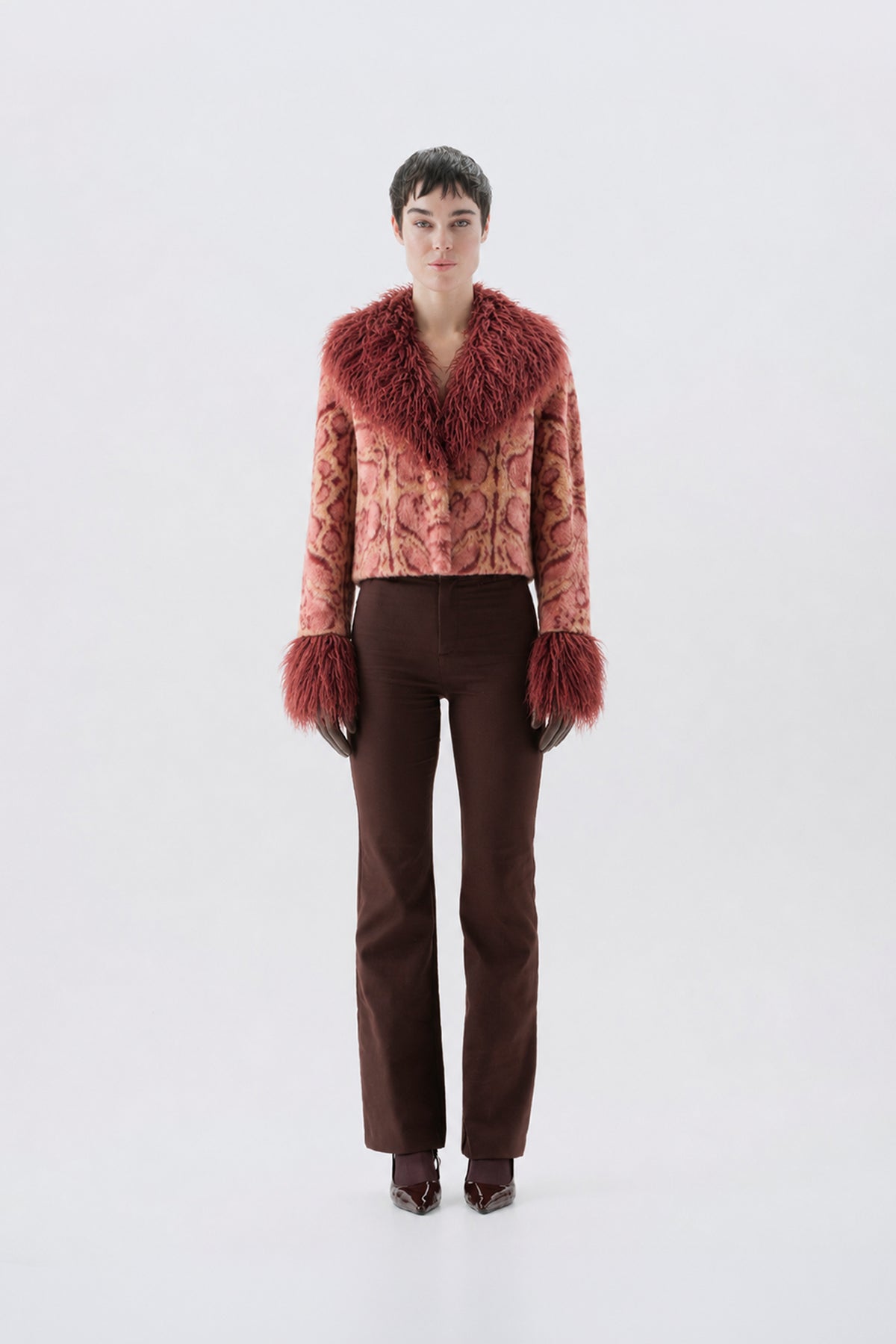 A model wearing a cropped pink patterned jacket with a large reddish-brown fur collar and matching fur-trimmed cuffs, paired with dark brown pants and dark shoes.