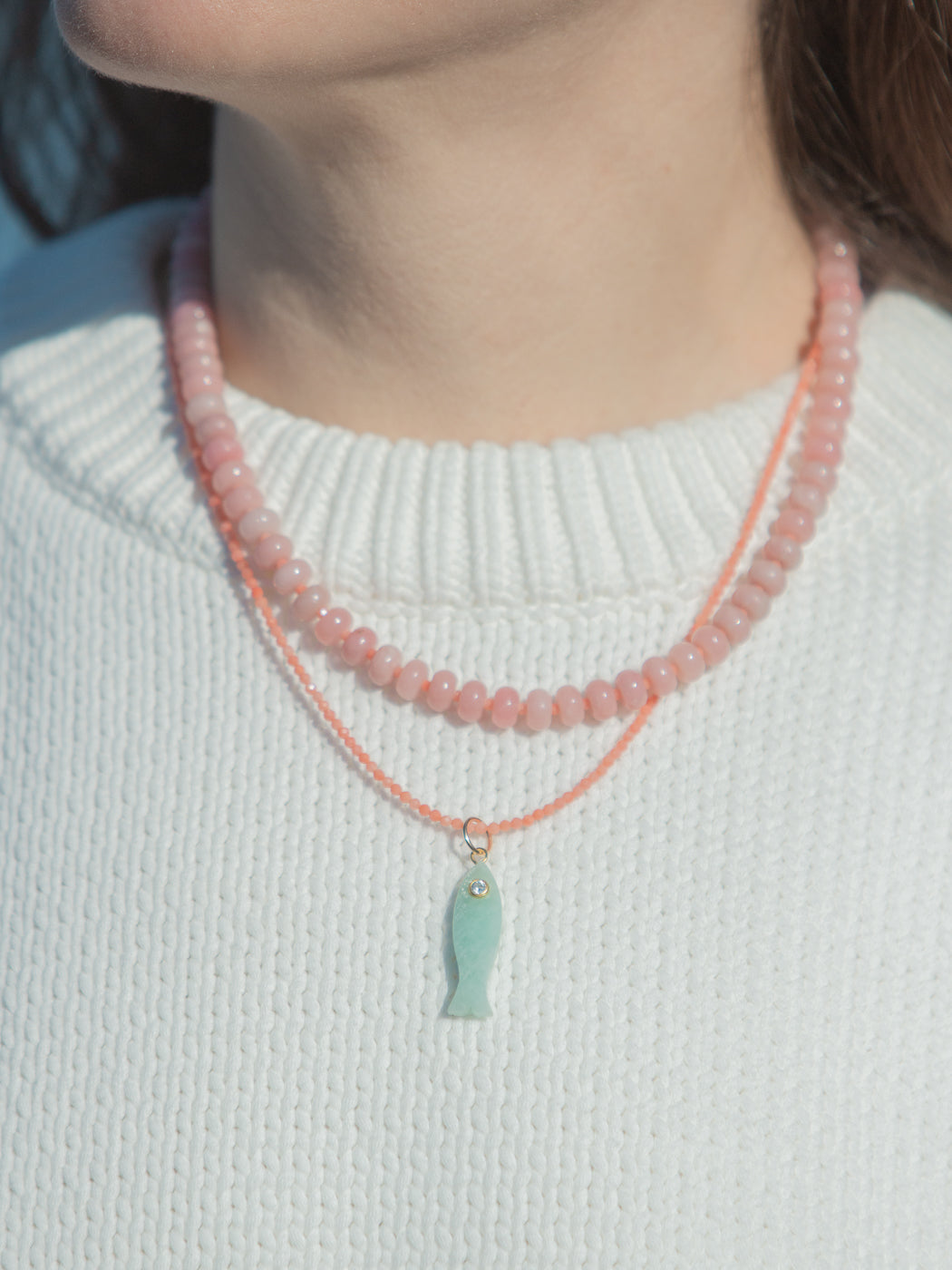 Close-up of Shay necklace in pink showcasing pink opal beads, coral-pink cord, and aqua fish pendant on white knit