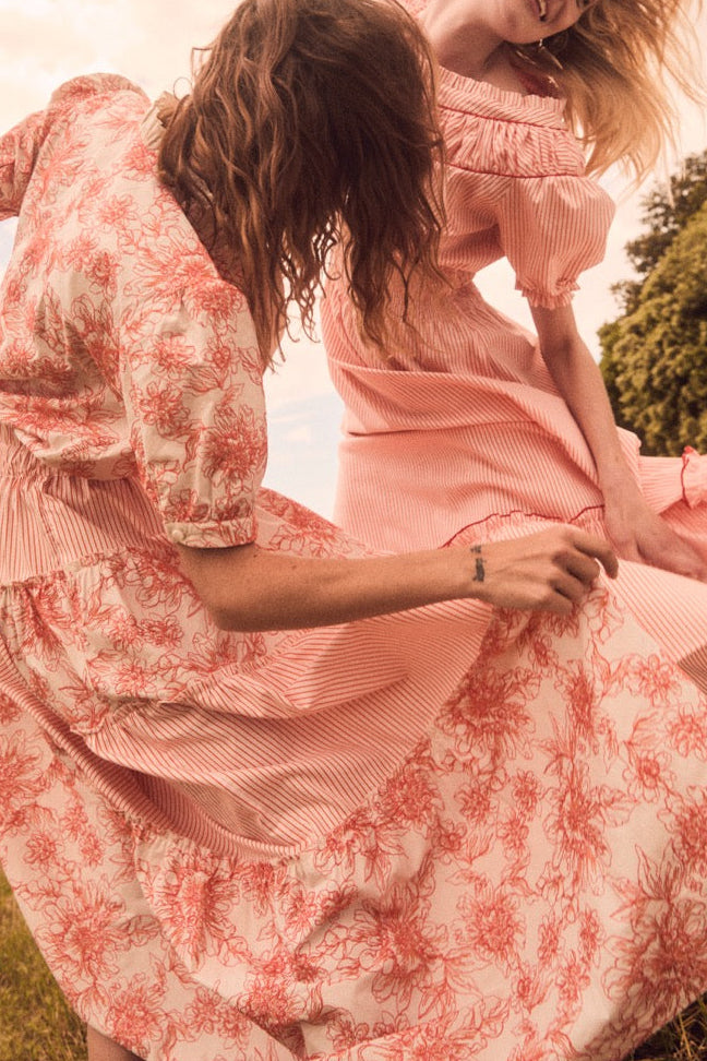 Two models twirling in a sunlit field wearing pink Dahlia Spectacle dresses, one wearing the Iole shirt with floral and striped smocking
