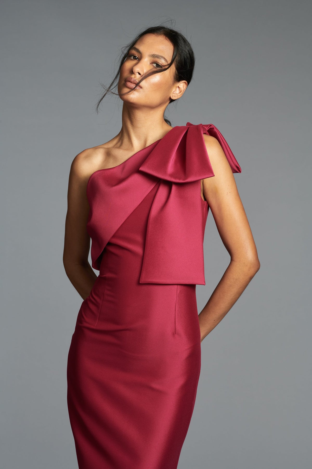 Front three-quarter view of Bonnie Gown in Rhodolite on model showcasing an asymmetric one-shoulder neckline with oversized bow and a column silhouette against a gray studio background