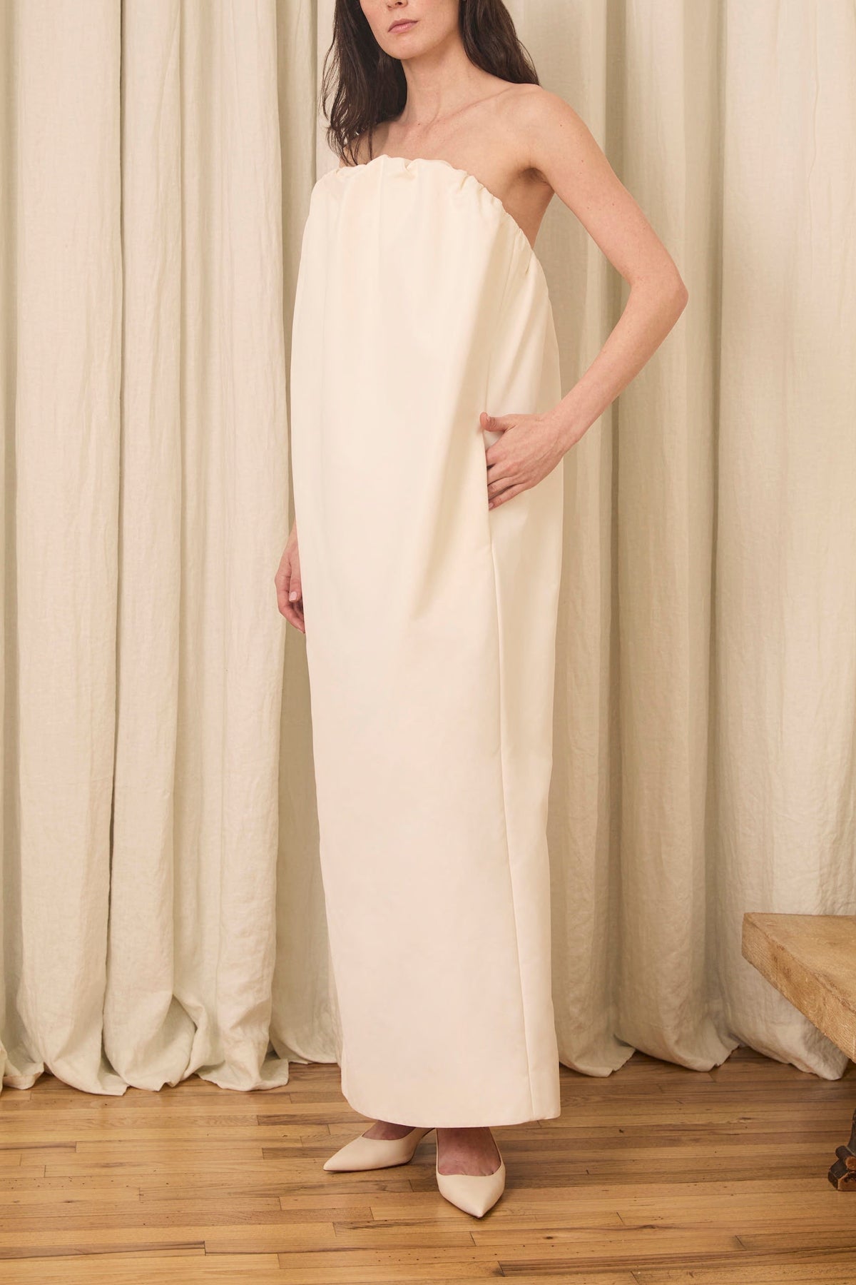 Side view of Simone Gown in Duchesse showcasing strapless gathered voluminous neckline and side seam pocket on model against cream curtain backdrop