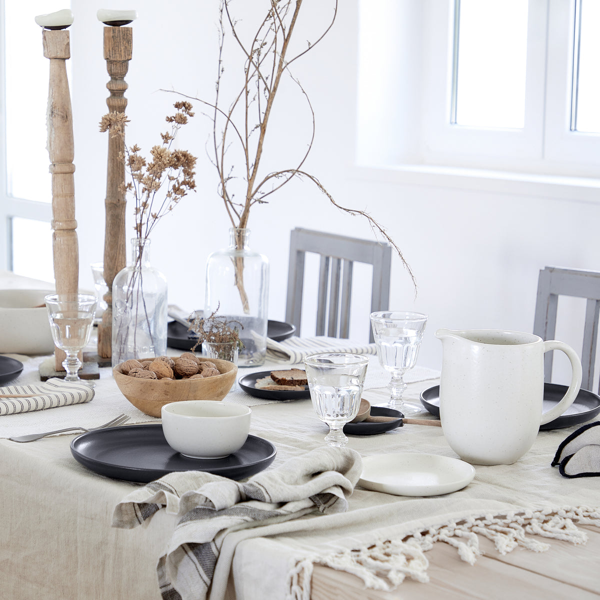 Sunlit dining table set with neutral stoneware plates and pitcher, glassware, wooden bowl of walnuts, dried floral centerpiece