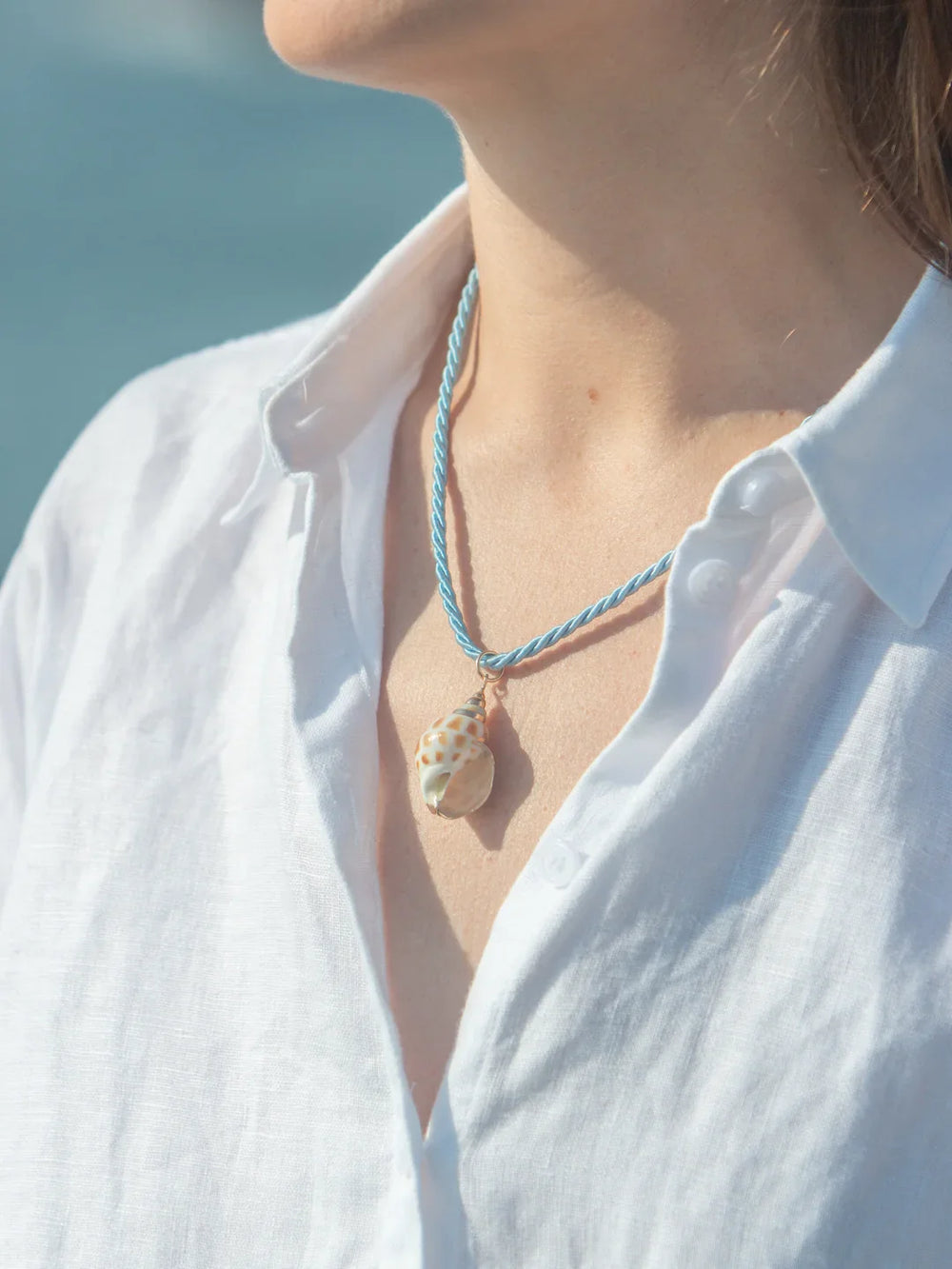 Close-up of Speckle Shell Charm necklace on model showing gold-plated speckled shell on a twisted blue cord against a white shirt