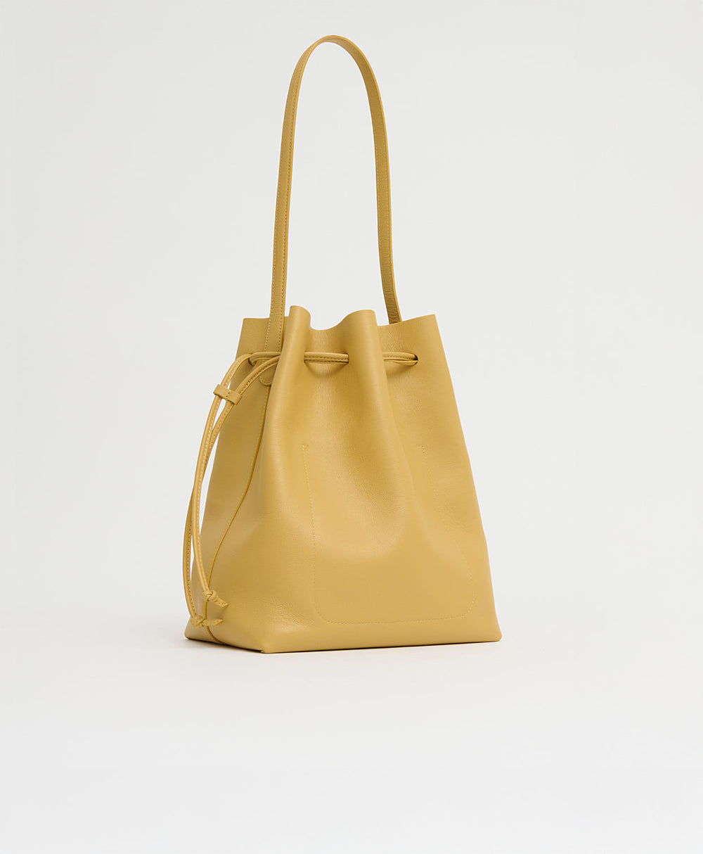 Three-quarter view of Drawstring Cabas Paglia showing drawstring closure, long shoulder strap, pebbled leather on white