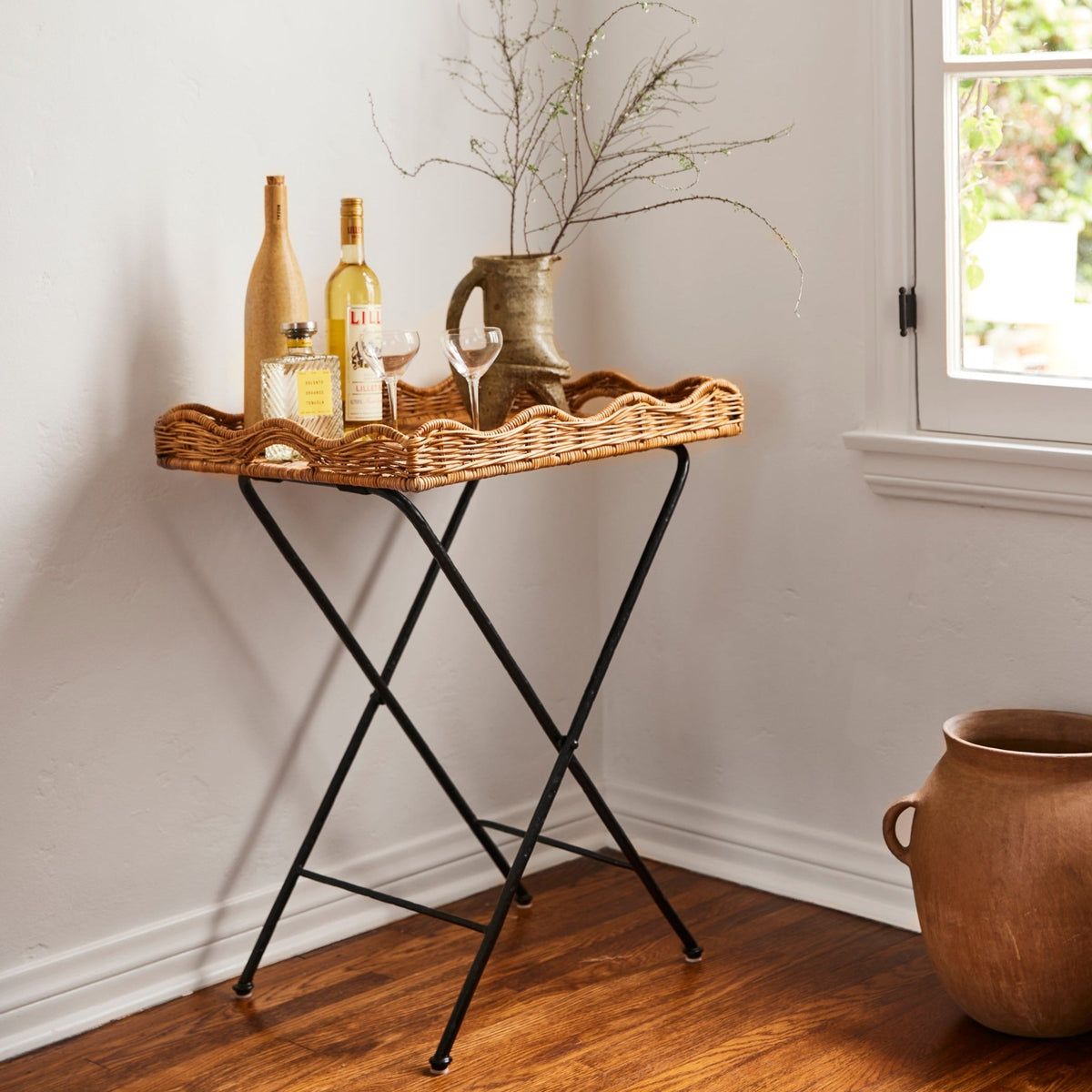 Angled view of Maude Scallop Butler Tray showing scalloped wicker tray on foldable black iron base with bottles and glasses