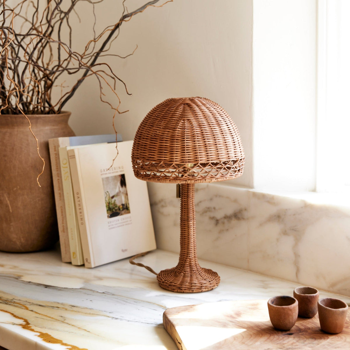 Front view of Augusta Mini Table Lamp showcasing woven rattan shade and stem on marble windowsill with books