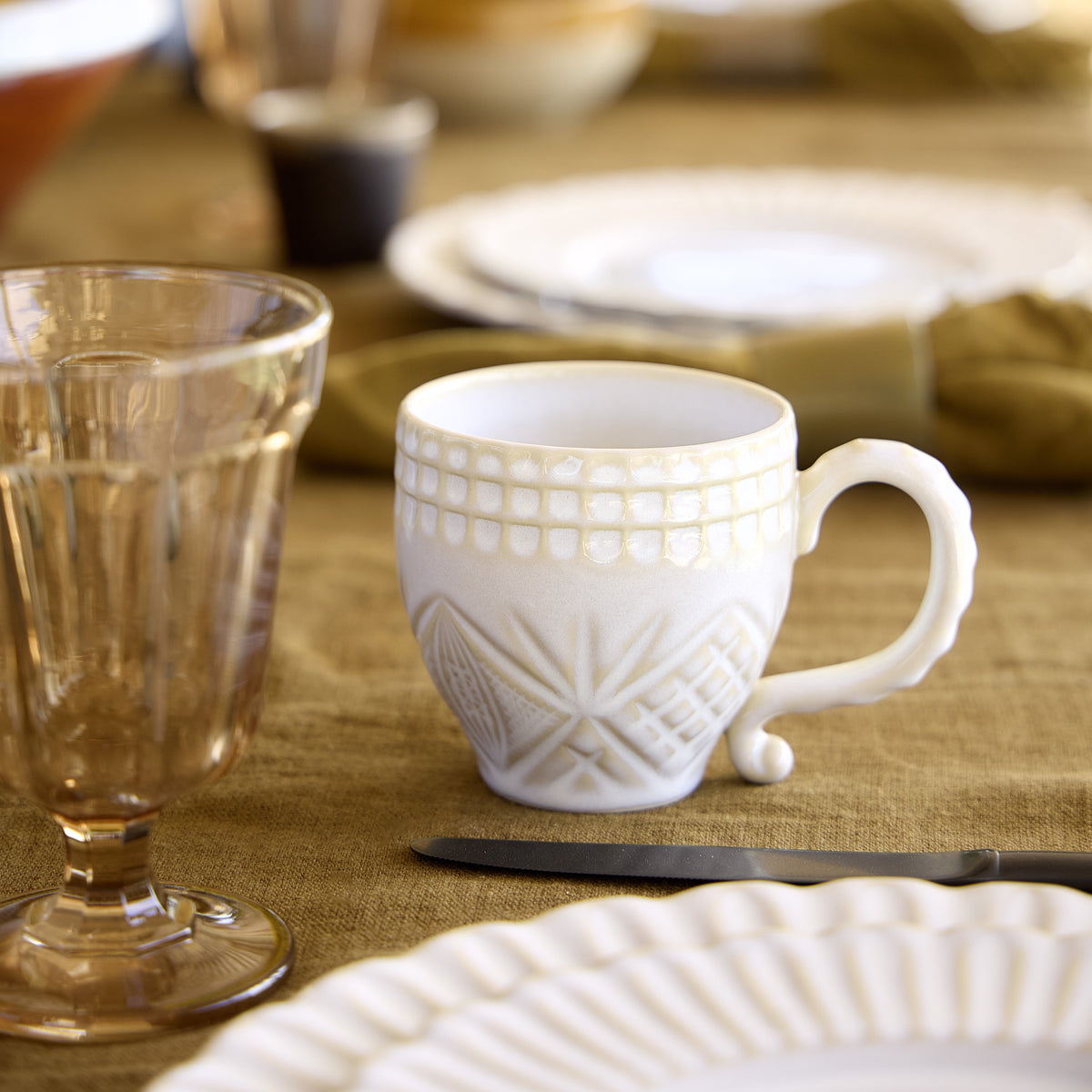 Front view of white Cristal Collection mug showcasing embossed crystal pattern and curled handle on a linen table setting