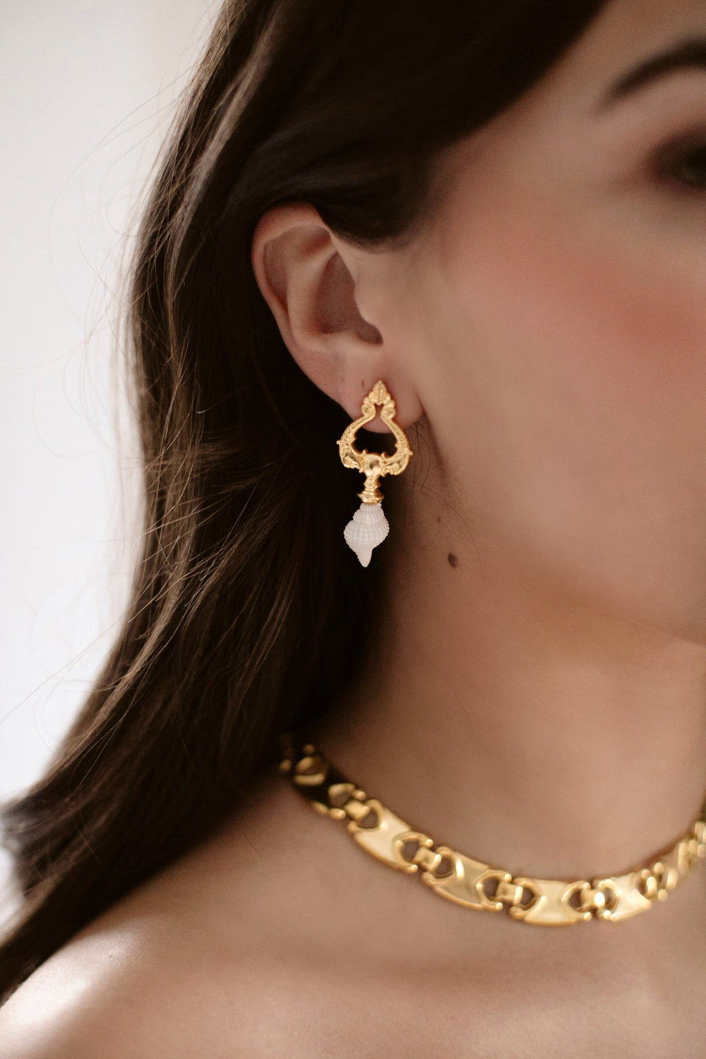 Close-up of Sadie Earrings on model ear showcasing 24k antique gold-plated dolphin topper and white trumpet shell drop on neutral background