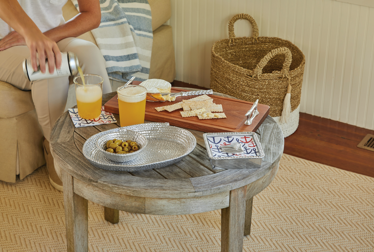 Rope oval platter holding olives on a rustic round coffee table in a living room, with a person pouring beer and a cheese board nearby