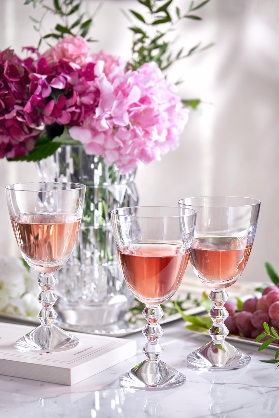 Front view of three Vega Water glasses showcasing stacked diamond-shaped stems with pink drinks and flowers in background