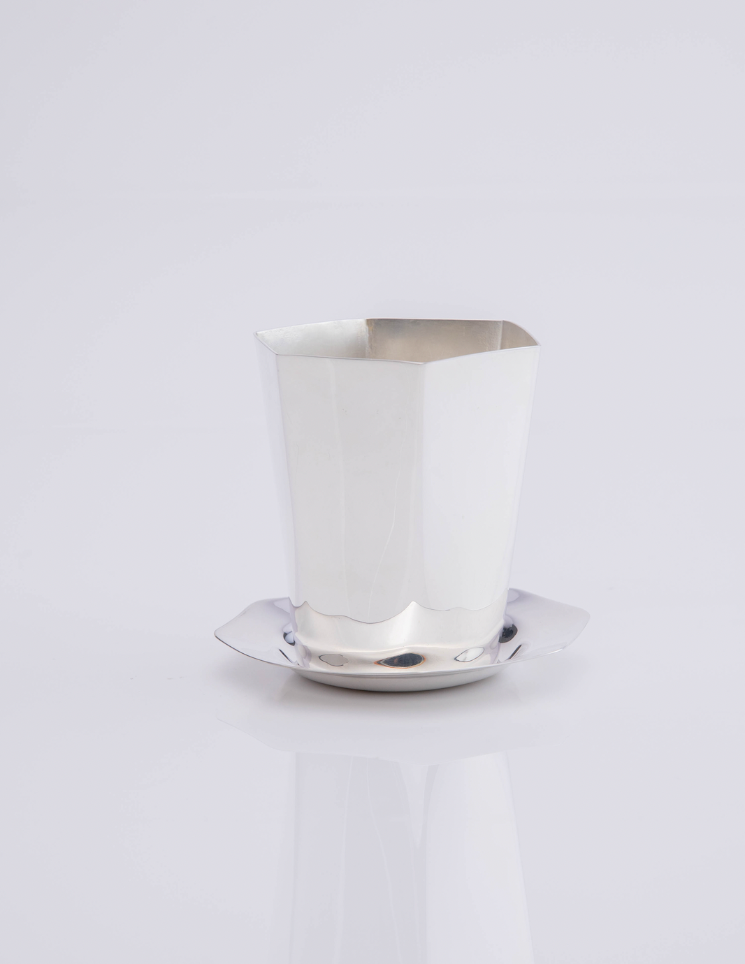 Front view of a hexagon coaster showcasing its silver-plated brass finish beneath a white faceted cup on a white background
