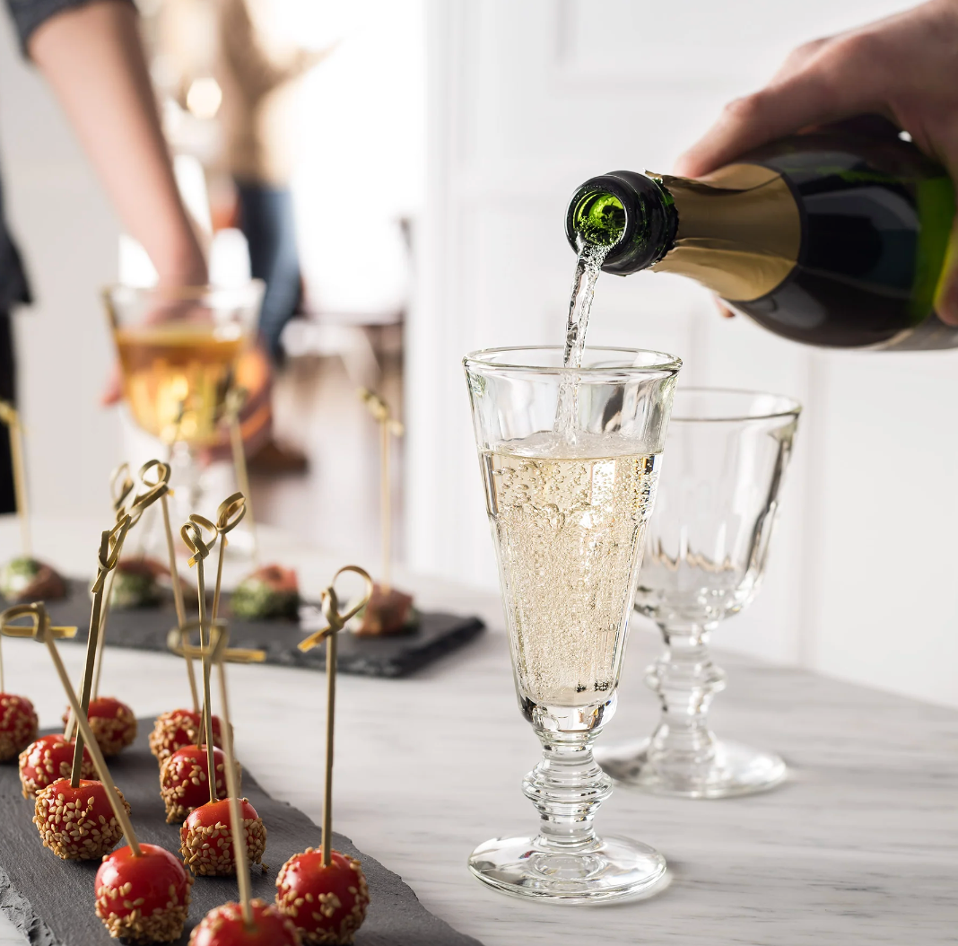 Front view of Perigold champagne flute with stacked stem as champagne is poured on a marble table with appetizers