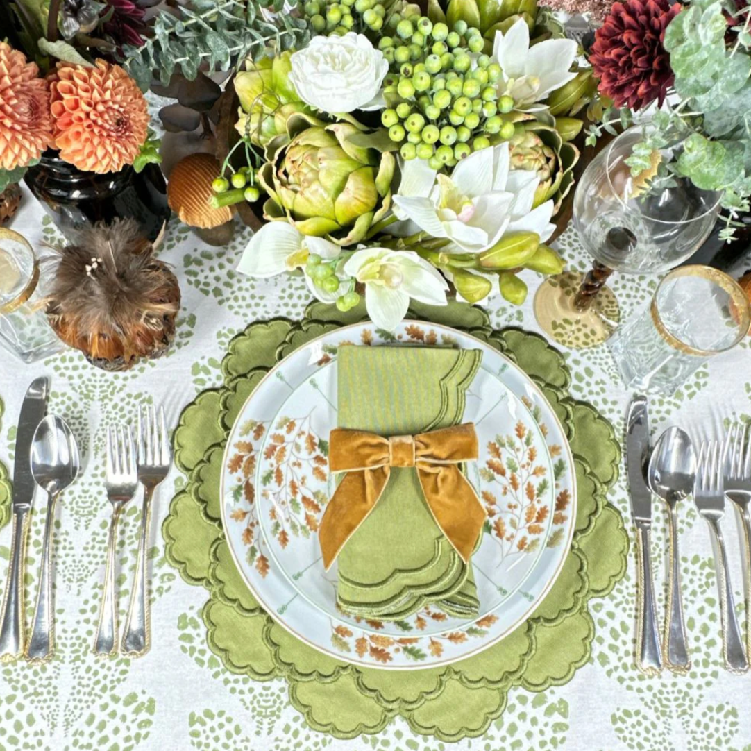 Top view of Lillian napkin in sage folded with a gold velvet bow on a scalloped sage placemat and floral table setting