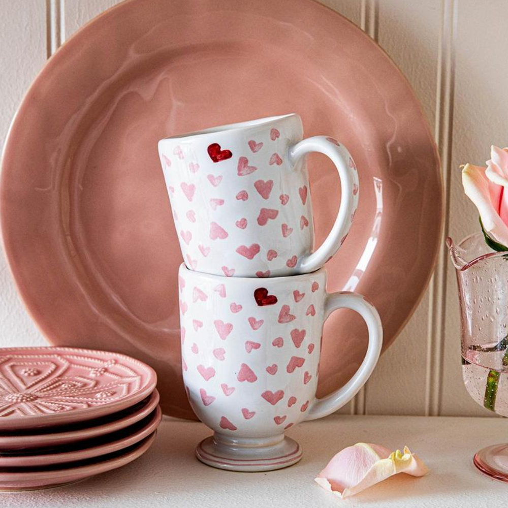 Front view of Love You More mug stacked to show pink heart pattern and pedestal foot against a pink plate background