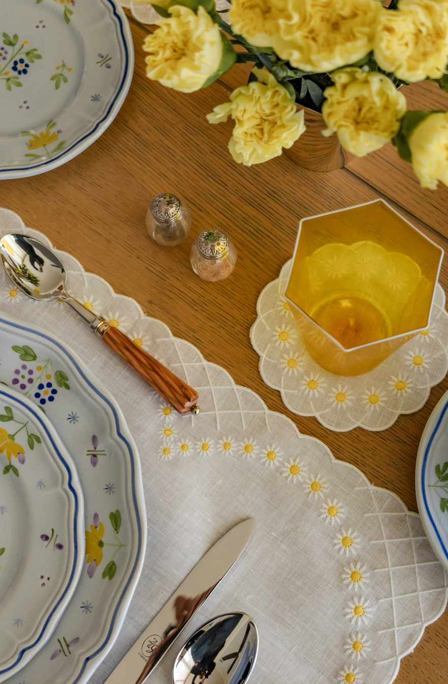 Overhead view of white Daisy coaster showcasing embroidered scalloped edge and daisy motifs on linen with plate and amber glass