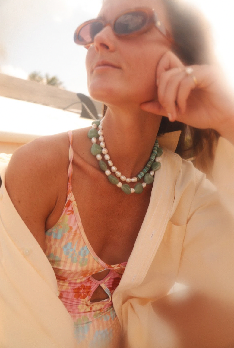 Woman wearing layered aqua heart stone and pearl necklaces while lounging outdoors in sunglasses and floral swimsuit
