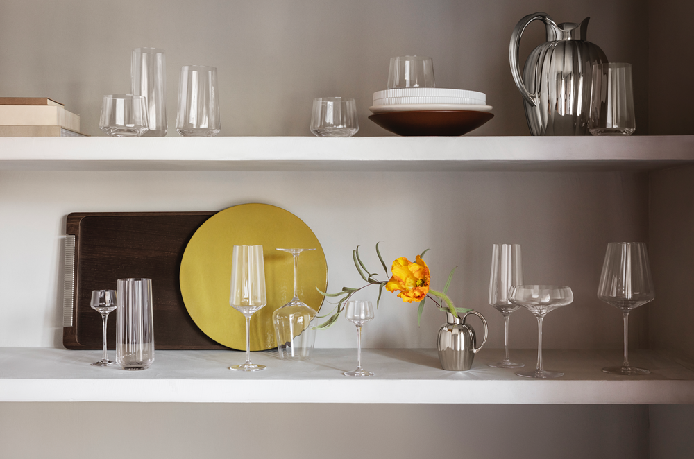 Bernadotte highball glasses displayed on white shelves with assorted glassware, a silver pitcher, yellow plate, and a single yellow flower