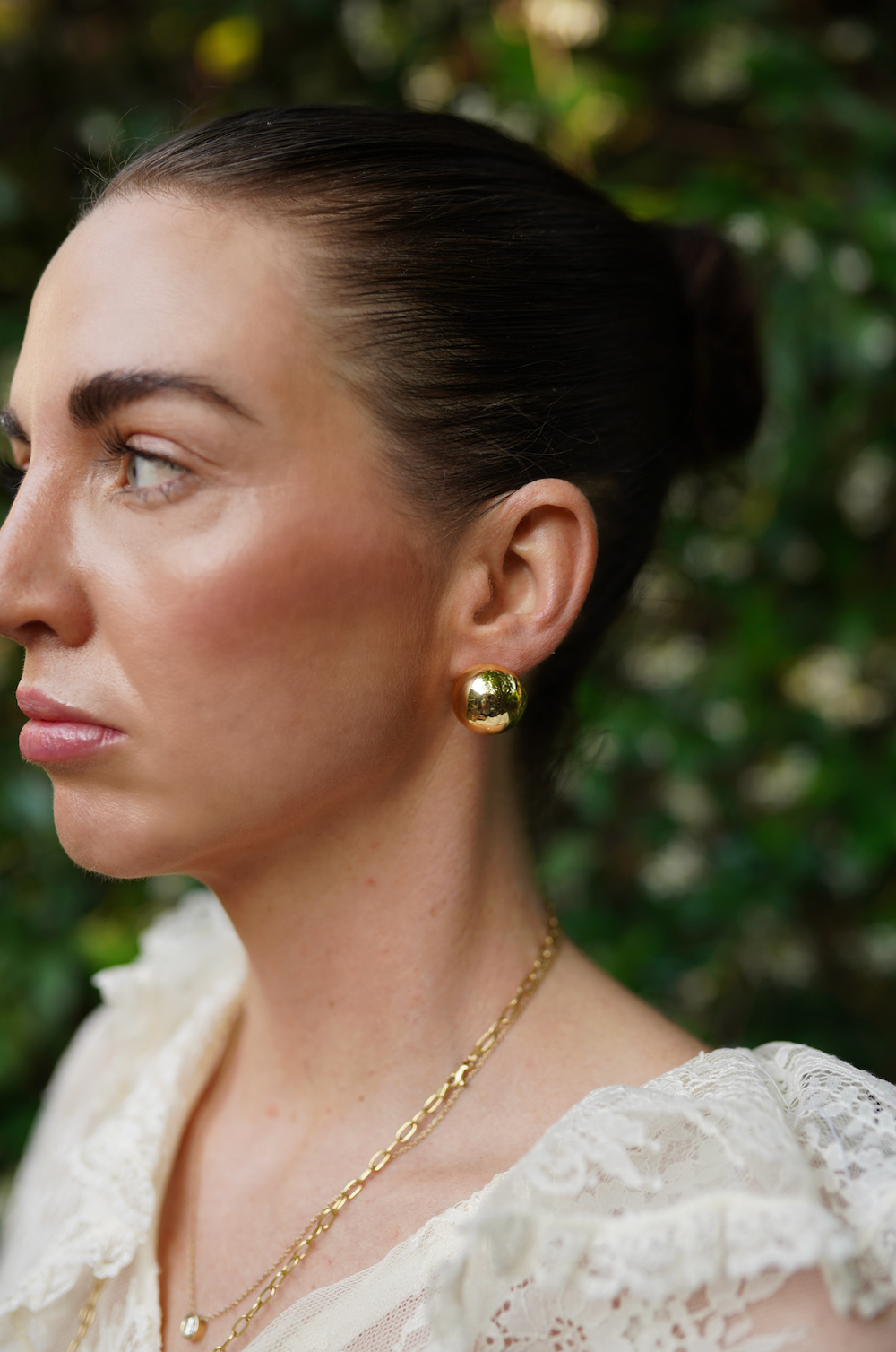Close-up left profile of model wearing The Cecilia gold-plated domed stud earrings against blurred green foliage background