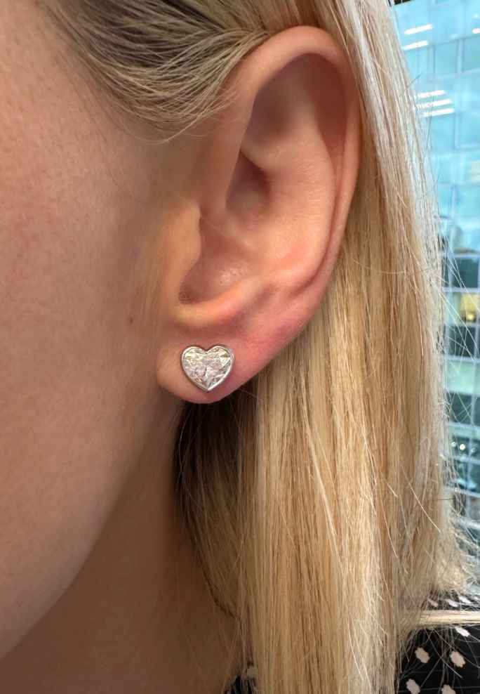 A close-up of a person’s ear wearing Bezel Set Diamond Heart Studs in 18k gold. Their straight, blonde hair frames the sparkling earrings, with a softly blurred glass building visible in the background.