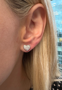 A close-up of a person’s ear wearing Bezel Set Diamond Heart Studs in 18k gold. Their straight, blonde hair frames the sparkling earrings, with a softly blurred glass building visible in the background.