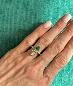A hand with light pink nails wears the Emerald and Diamond Double Shield Ring in Yellow Gold, featuring pear-shaped green and clear gemstones separated by a horizontal bar, set against a teal fabric background.