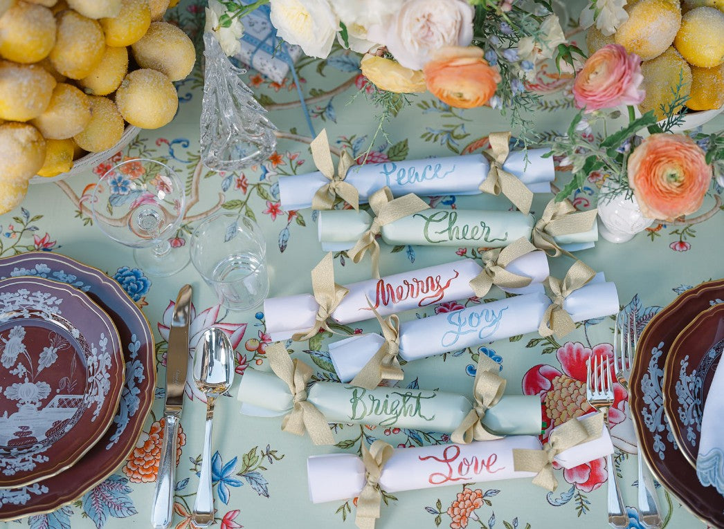 Top-down view of six pastel Over The Moon holiday crackers with gold bows and calligraphy reading Merry, Peace, Cheer, Joy on a floral tablecloth