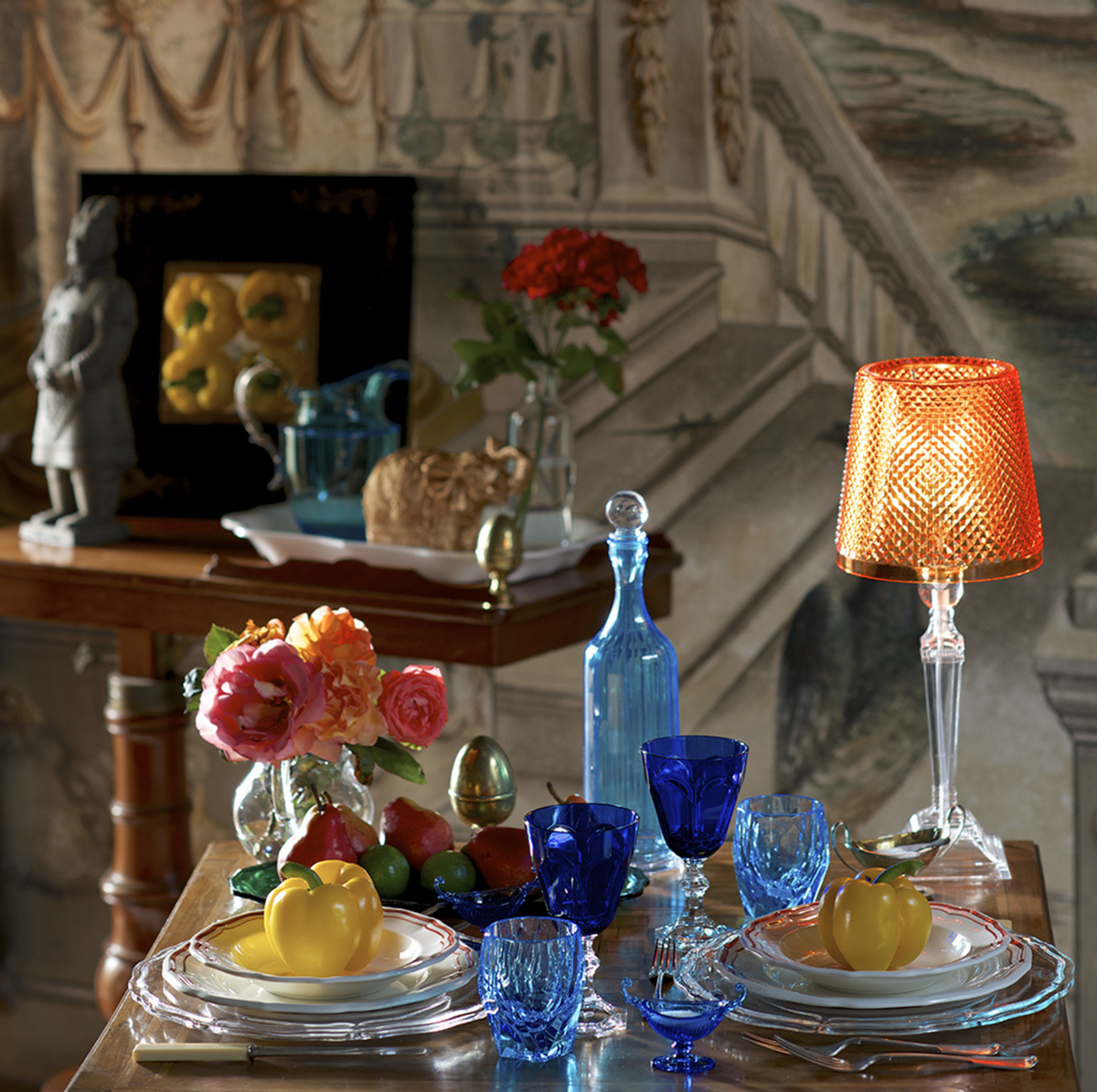 Table set for two with blue glassware, stacked clear plates and two yellow Milly large acrylic tumblers, roses and an amber lamp in the background