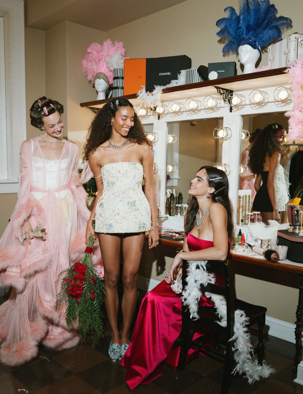 Three women backstage, seated woman in fuchsia wearing tourmaline and diamond necklace while friends in pastel robes smile