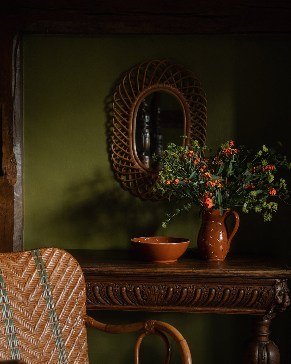 Front view of oval rattan mirror showcasing woven looped frame hanging on an olive-green wall above a carved wooden console