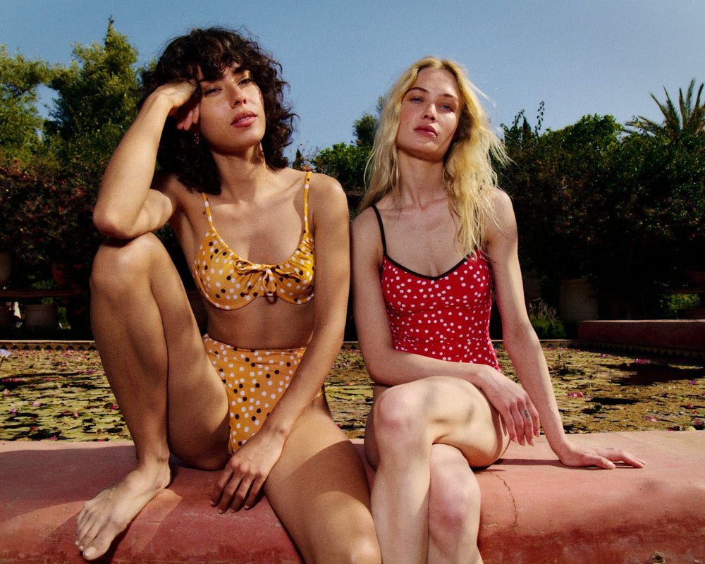 Two models seated by a garden pond wearing The Rose bottom in Marigold Ikat Dot and a red polka-dot one-piece