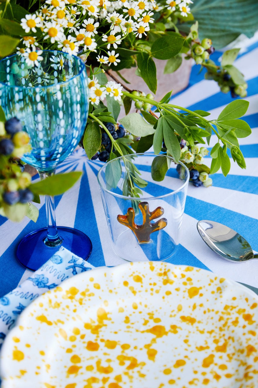 Blue Perle wine goblet next to a clear tumbler and yellow-splattered plate on a blue-and-white striped tablecloth with daisies