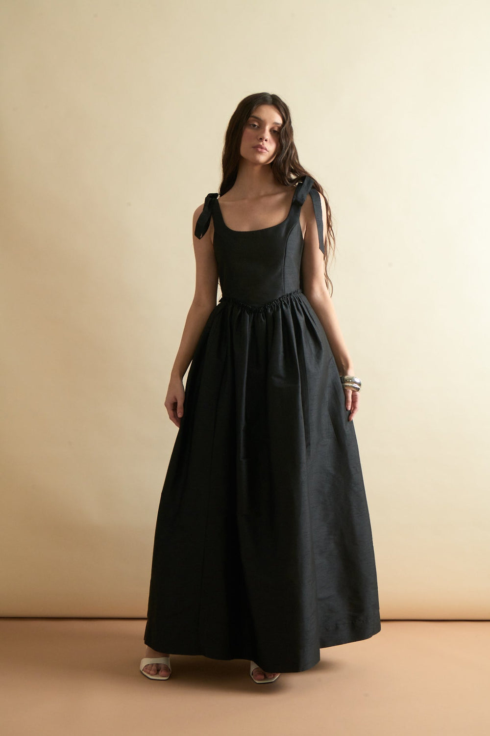 Front view of Marie Dress in black on model showcasing scooped bodice, tied shoulder straps, and full gathered skirt against a neutral beige background