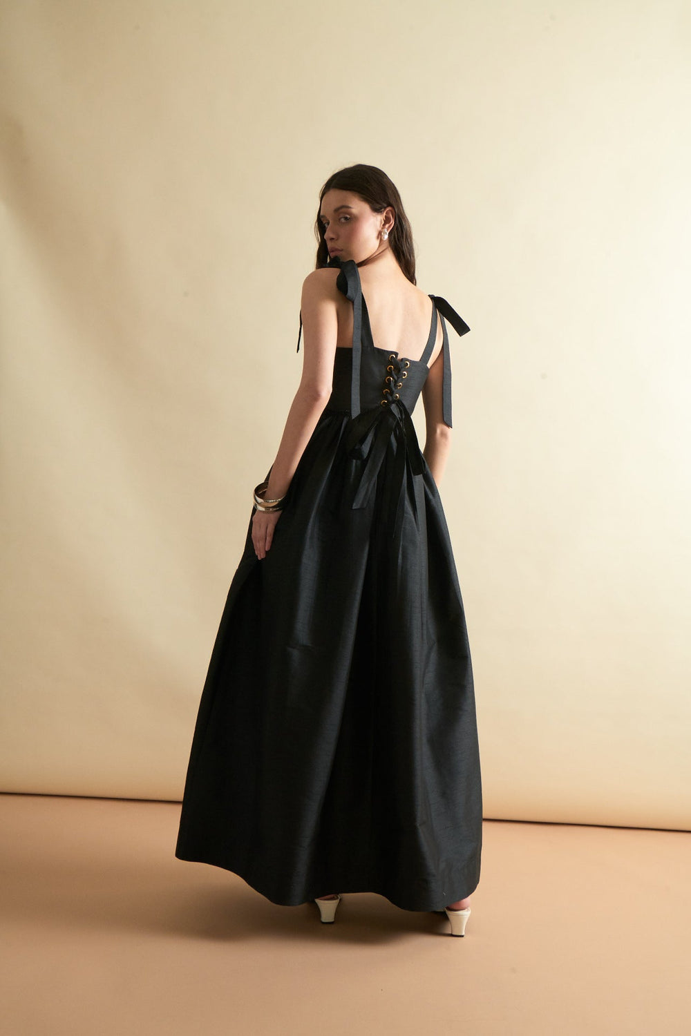 Rear view of Marie Dress in Black showing lace-up corset back, tied shoulder straps, and full floor-length skirt on beige background