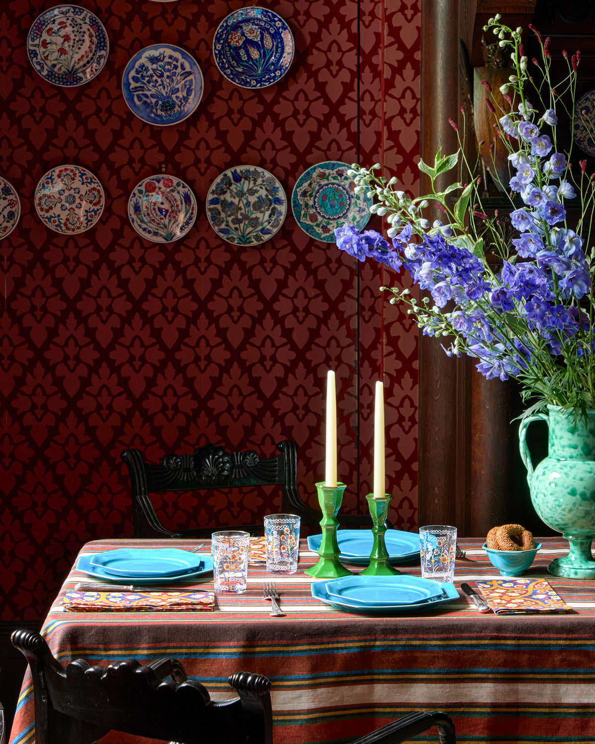 Striped linen tablecloth covers a dining table set with blue plates, patterned napkins, glasses, and green candlesticks