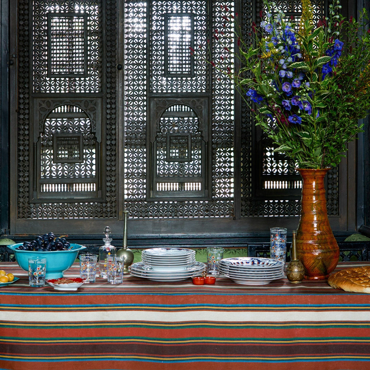 Front view of Tingere Natural Striped Tablecloth, Square showcasing deep brown, red, and white stripes with tableware and flowers