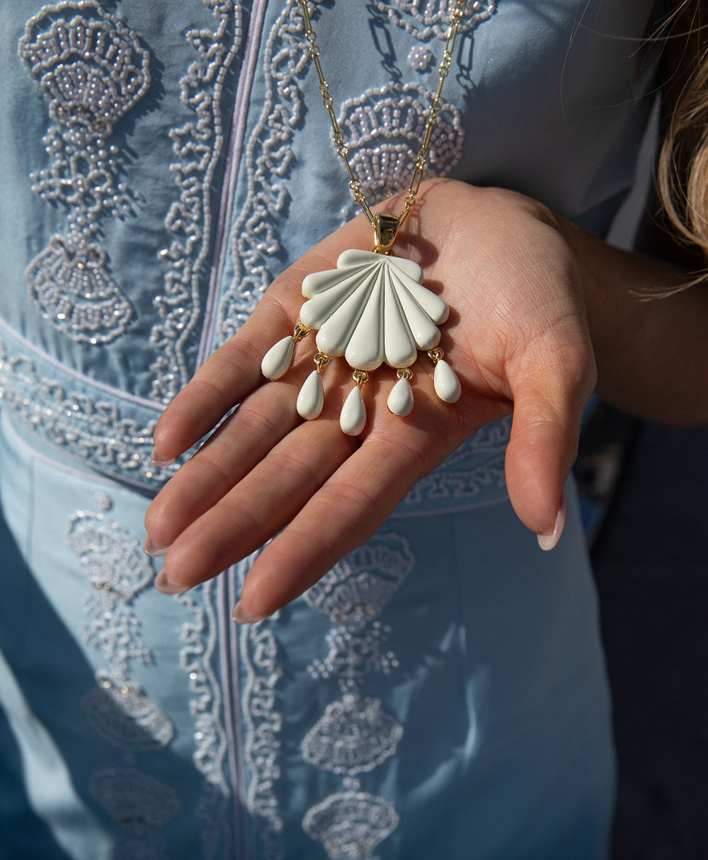 Close-up of Sirena pendant, ivory resin shell with gold bail and teardrop dangles, shown in hand over embroidered blue dress