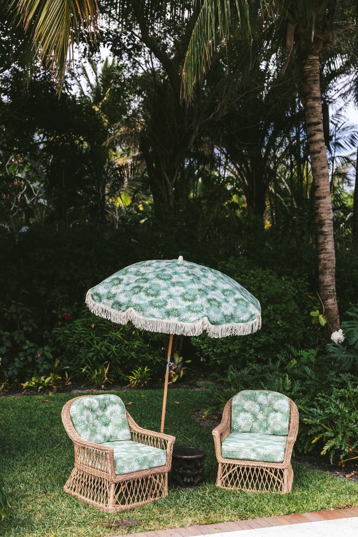 Two wicker patio chairs with green palm-print cushions under a matching fringed umbrella on a lawn in a tropical garden